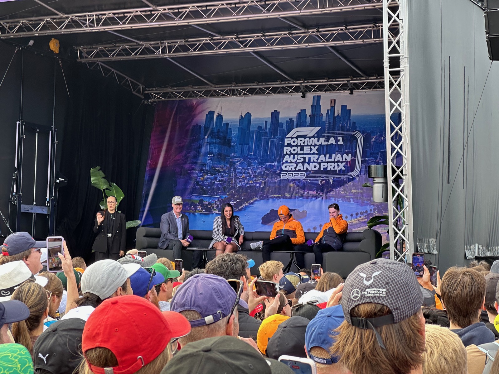 Four people on stage, two in orange, two in suits, facing audience with cityscape backdrop.