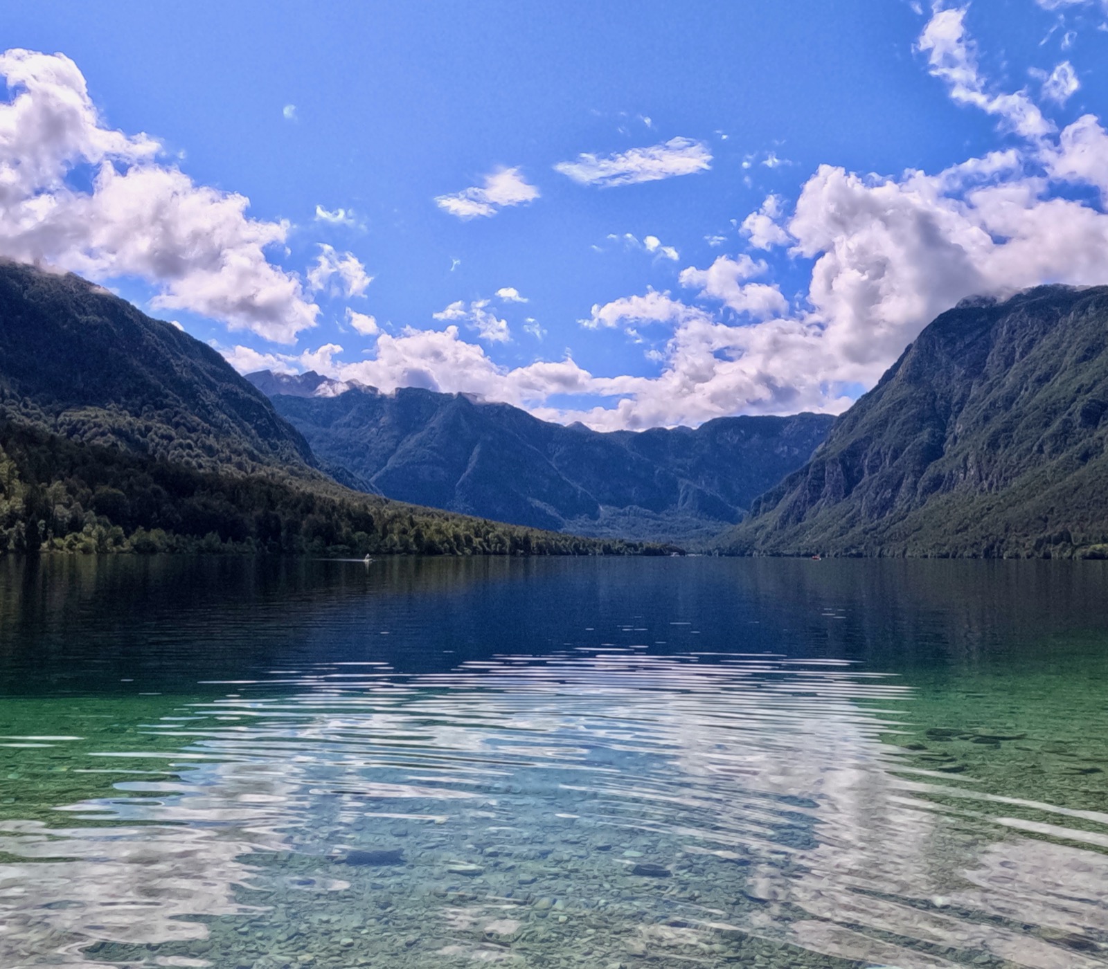 Clear blue lake with rocky bottom, surrounded by forested mountains under a partly cloudy sky.