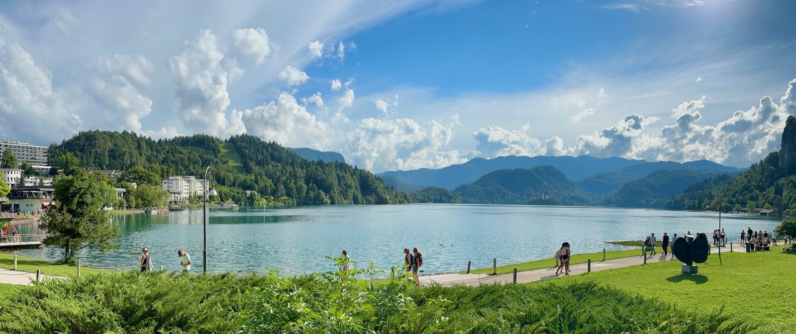 People walking on a path next to a lake with mountains and buildings in the background.