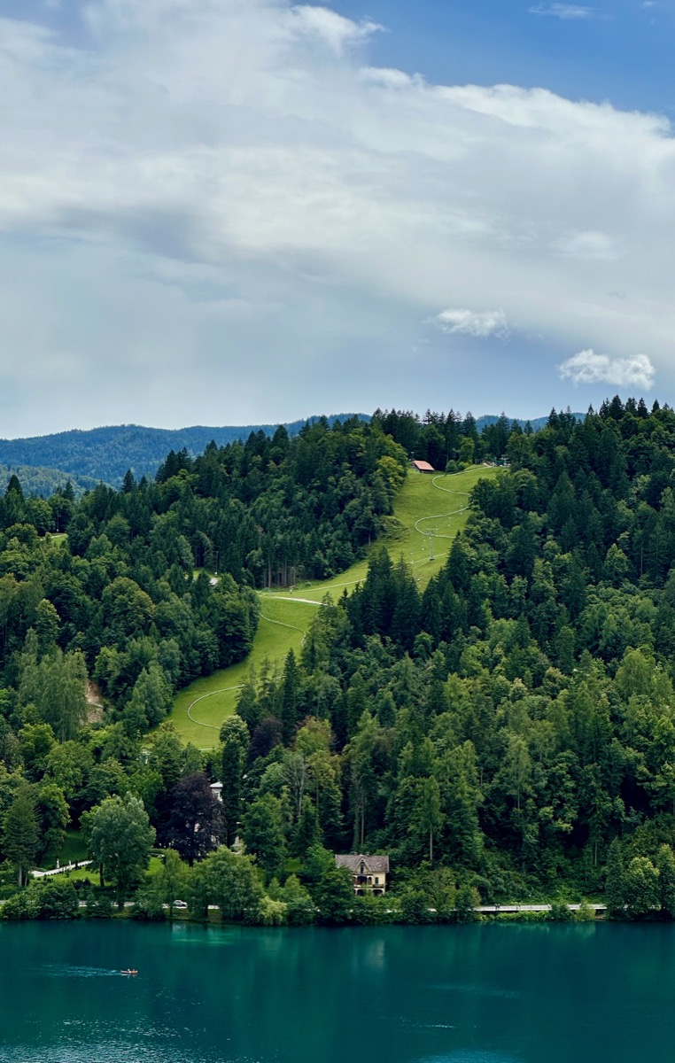 Green hill with trees, red roof, and lake with boat under blue sky.