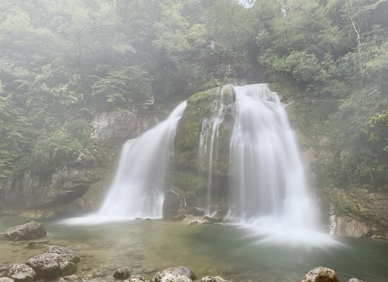 Two waterfalls flowing into a pool surrounded by rocks and trees.