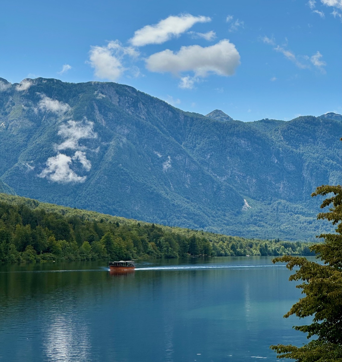 Red boat on calm lake with forested shore and mountains under blue sky.