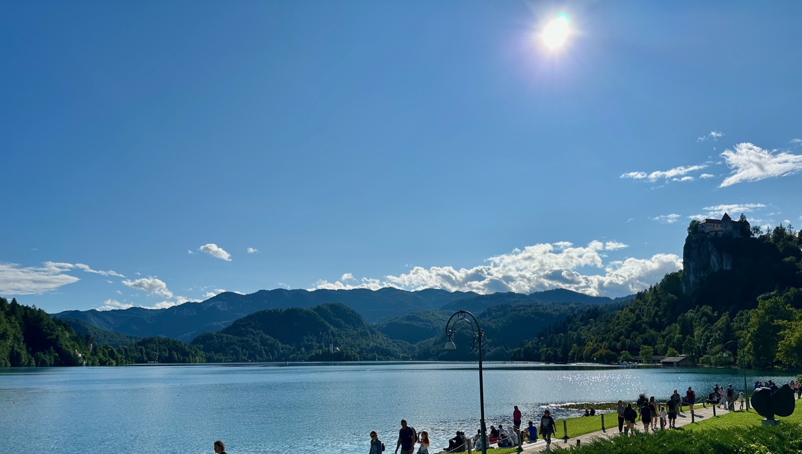 People walking on a path next to a lake with mountains and a castle in the background.