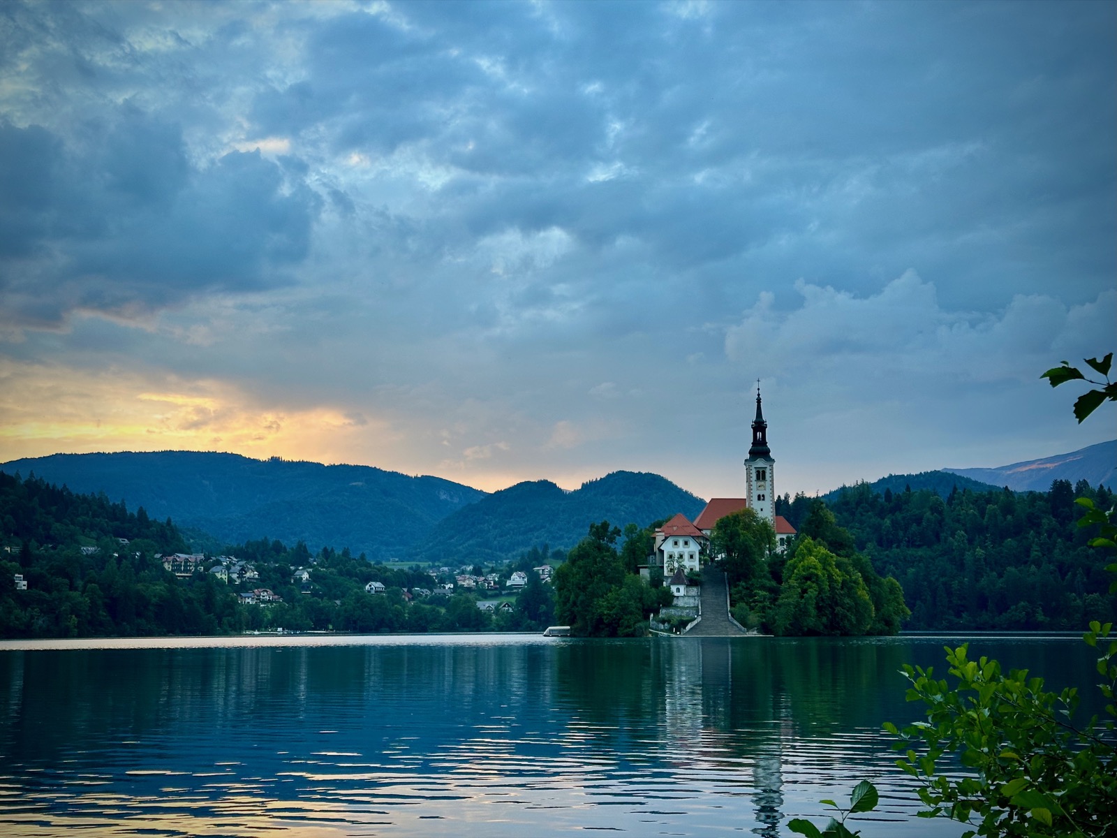 Church on island with red roof, surrounded by water and trees, under cloudy sky.