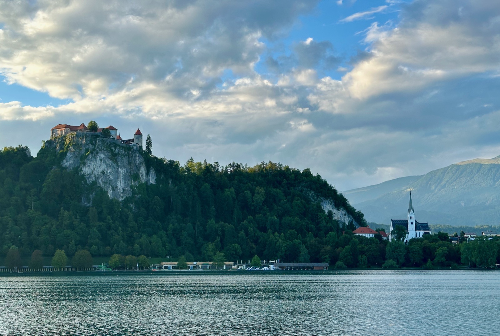 Castle on rocky cliff above lake with church and mountains.