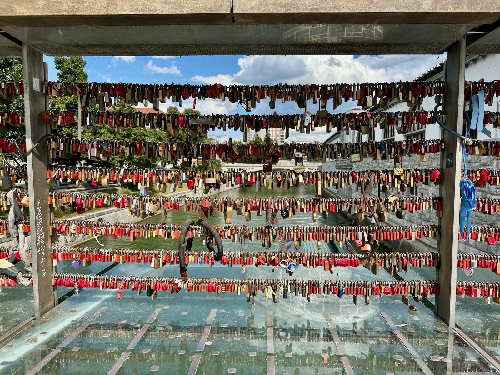 Red and black padlocks hang from metal bars over a river with stone bridges and buildings in the background.