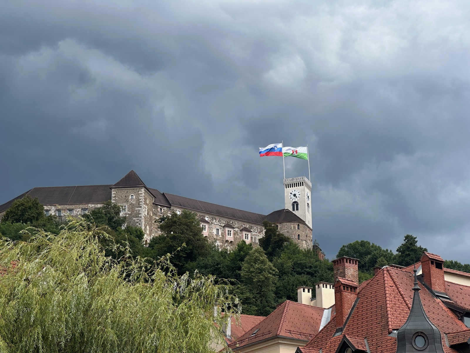 Two flags on a tower above a stone building with red roofs and trees.
