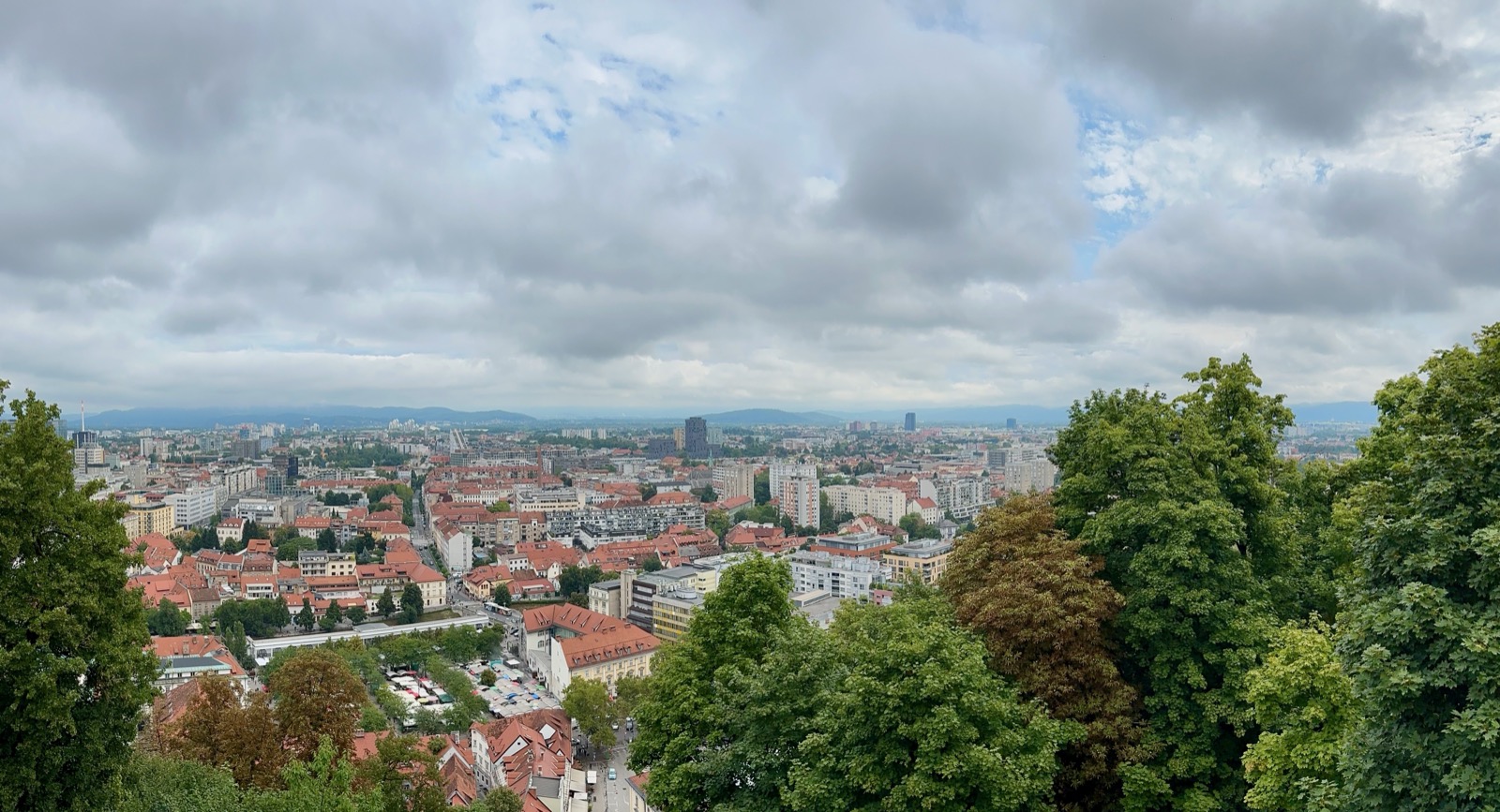 Trees frame a cityscape with red-roofed buildings and distant mountains under a cloudy sky.