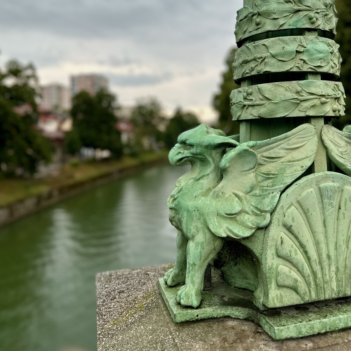Green dragon statue with wings on stone bridge railing, river and buildings in background.