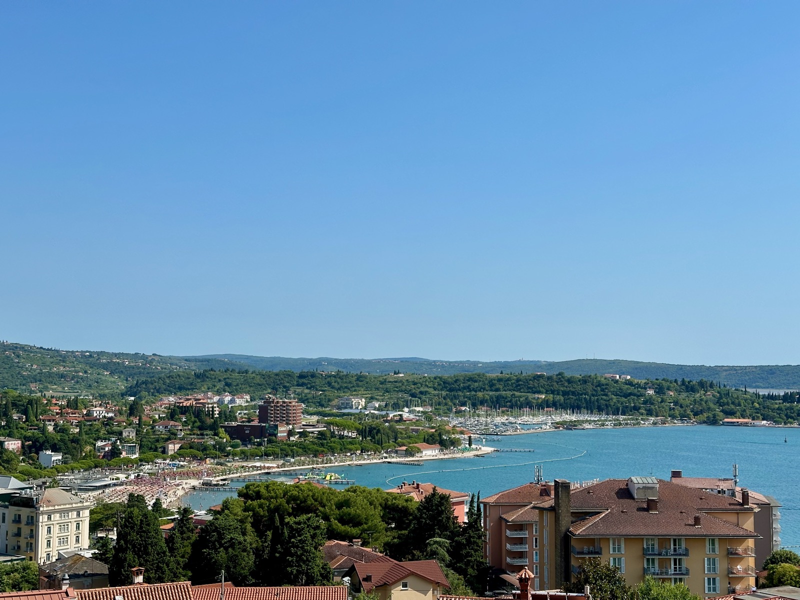 A coastal town with buildings, trees, and a marina under a blue sky.