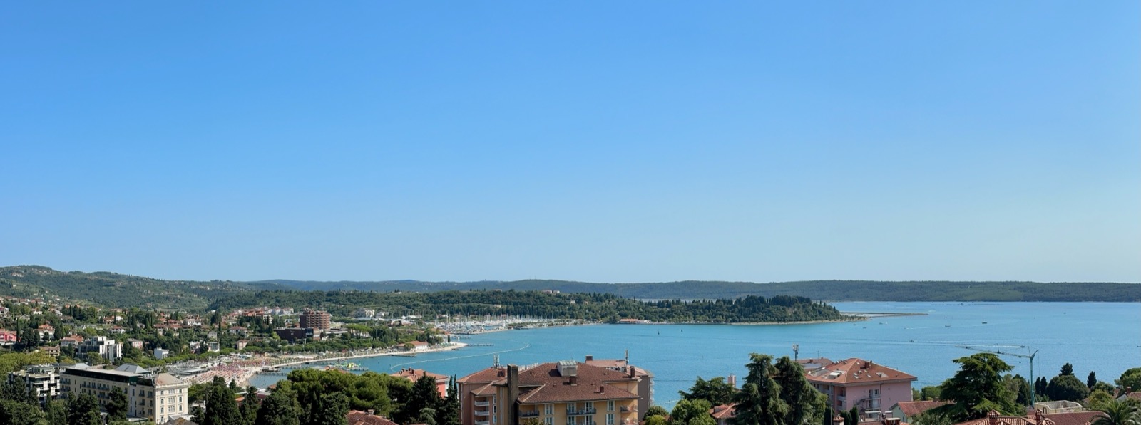 Blue sea, green hills, buildings, and boats under a blue sky.