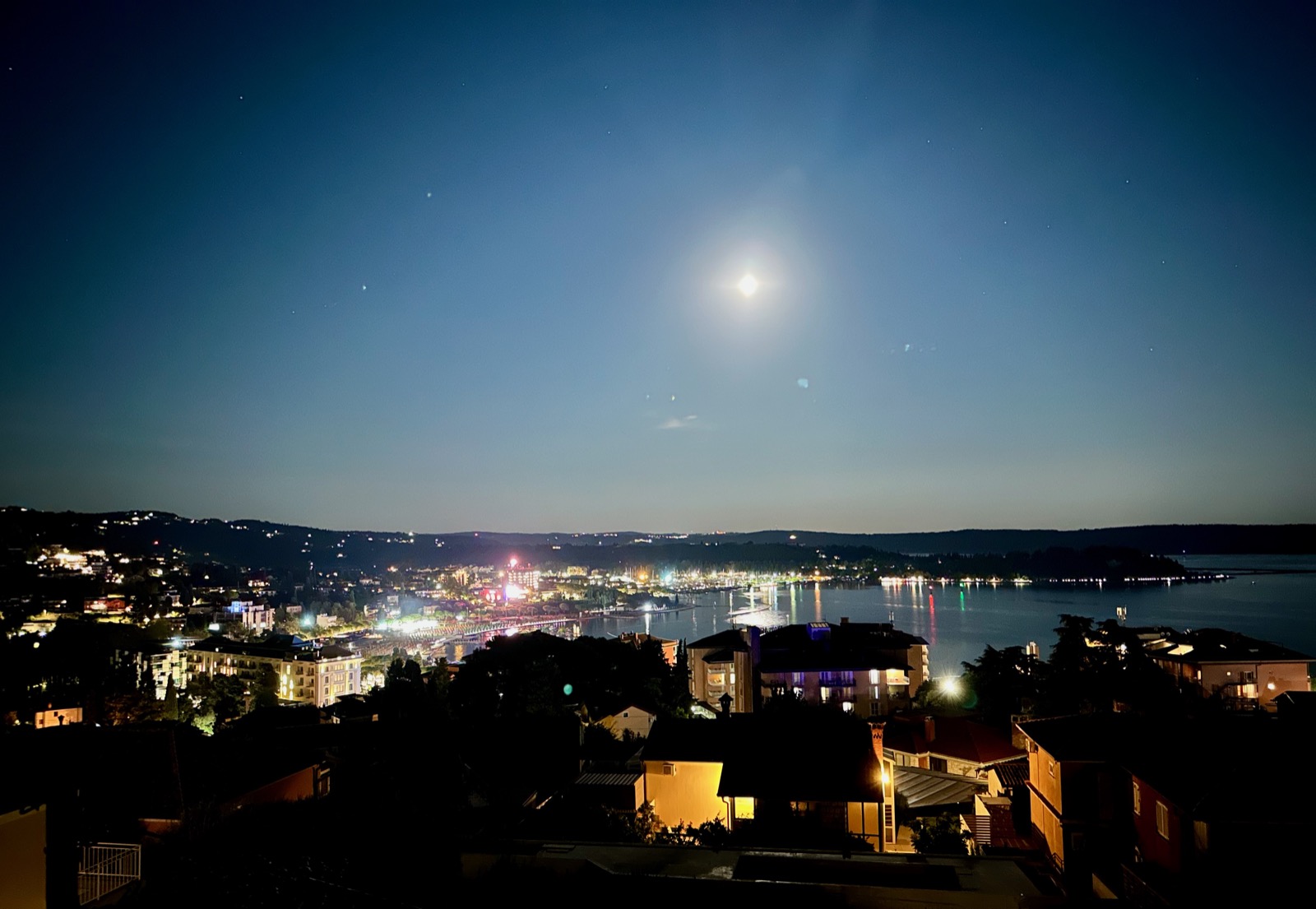 Cityscape with buildings, lights, and water under a moonlit sky.