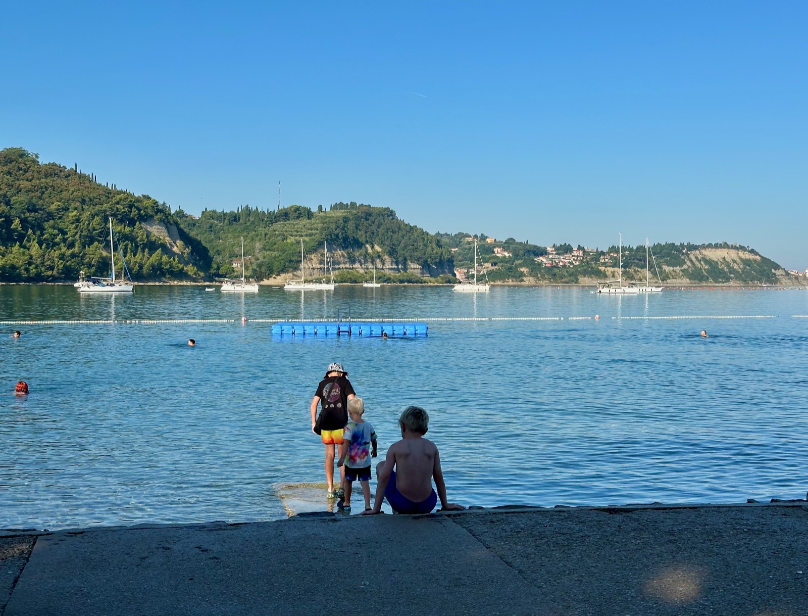 Three children sitting on a concrete ledge overlooking sailboats and a blue floating platform in Portoroz, Slovenia.