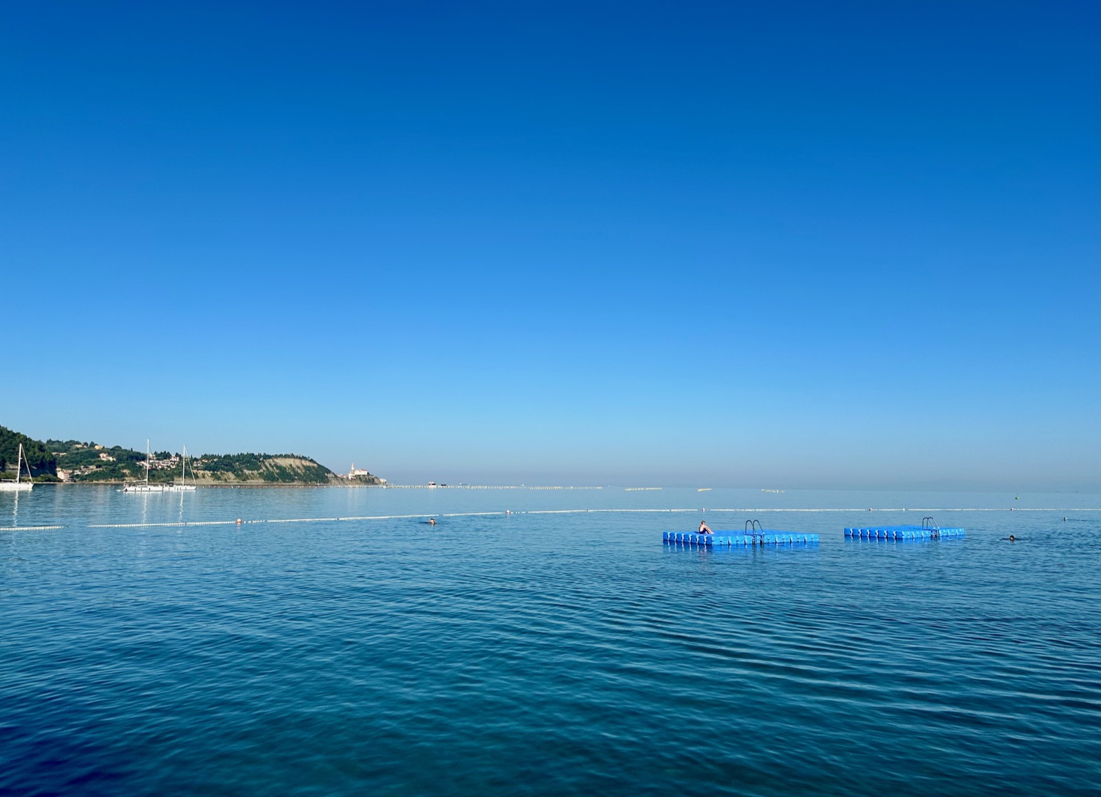 Blue floating pool with ladders in calm sea near coastline with buildings and trees.