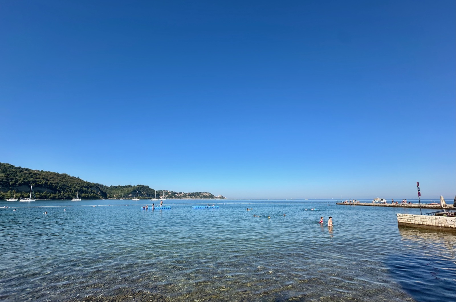 People swimming and standing in shallow water near a pier under a blue sky.