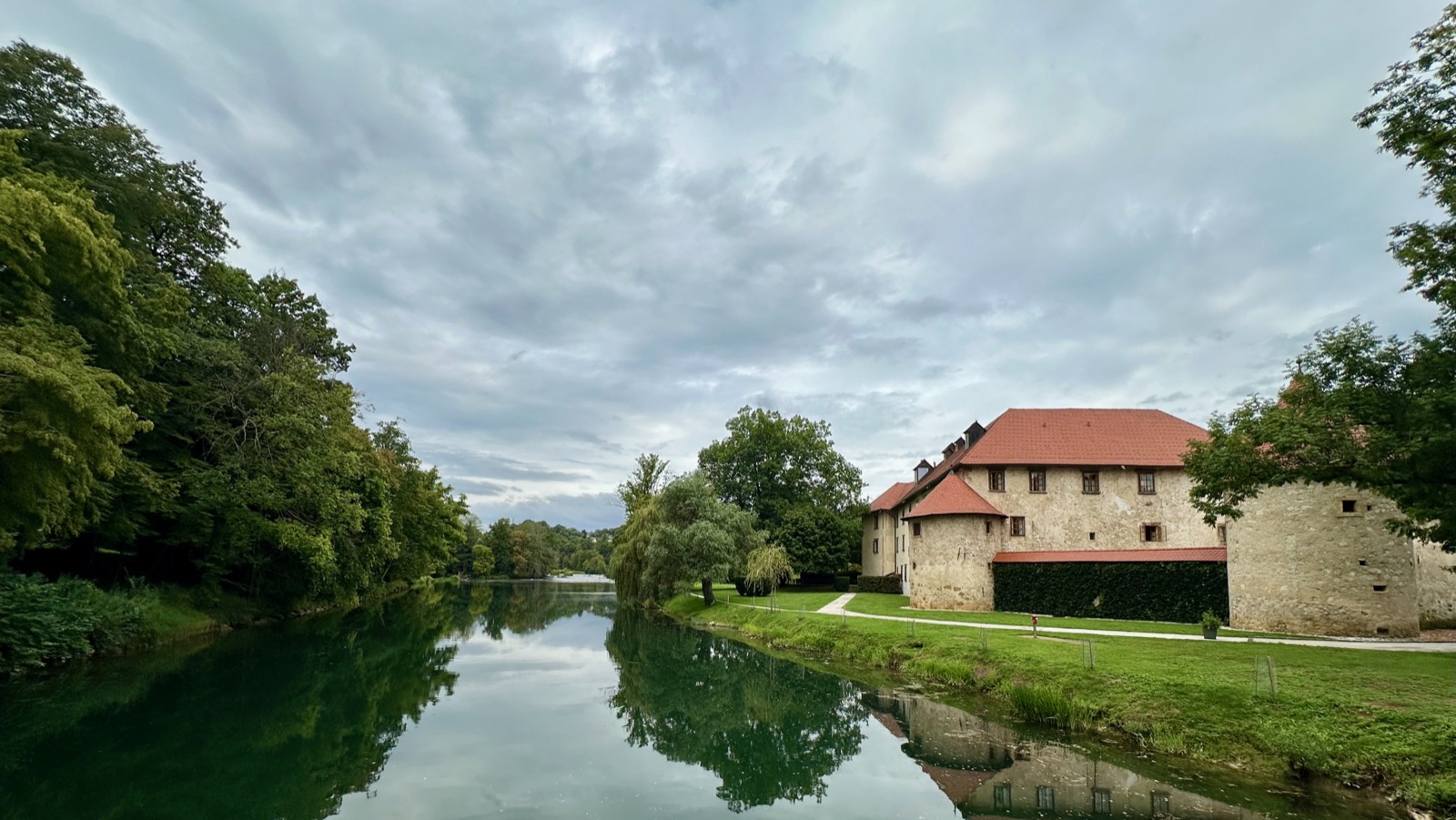 Castle with red roof by river, surrounded by trees and grass.